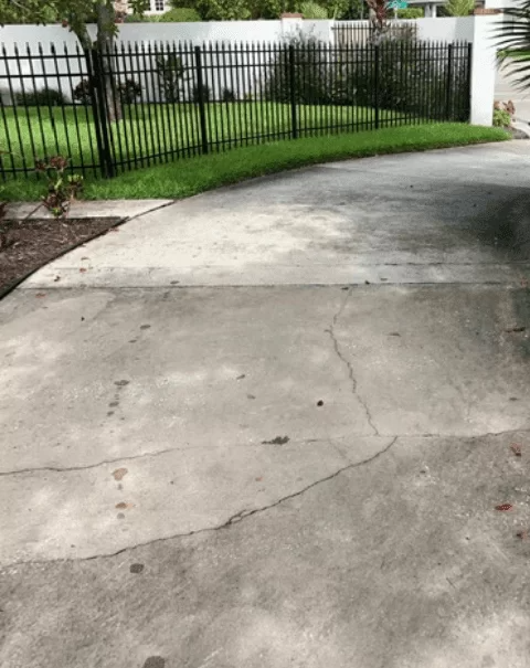 Cracked concrete driveway curving past a black metal fence and green lawn