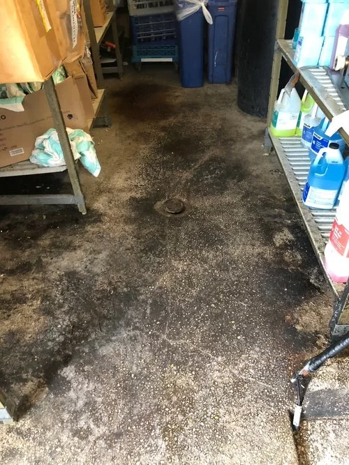 Dirty, stained concrete floor in a narrow storage room with shelves and boxes along both sides.