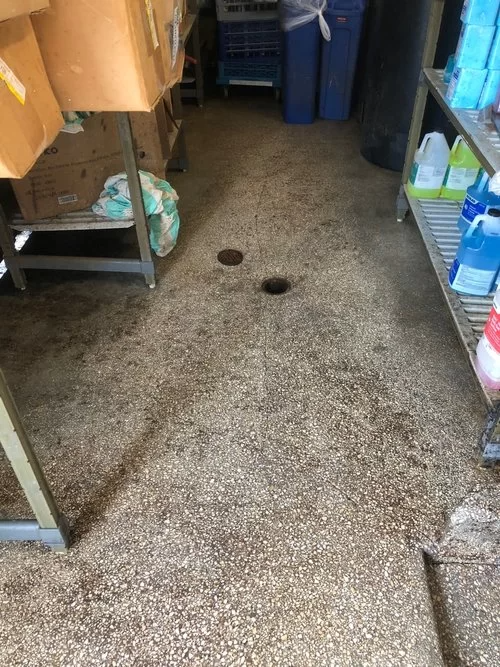 A narrow storage aisle with shelves, cardboard boxes, blue bins, and a concrete floor with two holes.