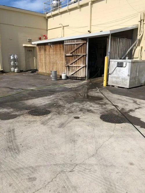 Outdoor service area with concrete ground, open utility door, wooden barrier, and trash bin beside a beige building