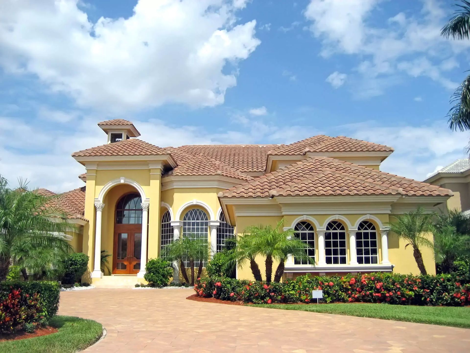 Yellow Mediterranean-style house with terracotta roof, arched windows, and a landscaped front garden under blue sky