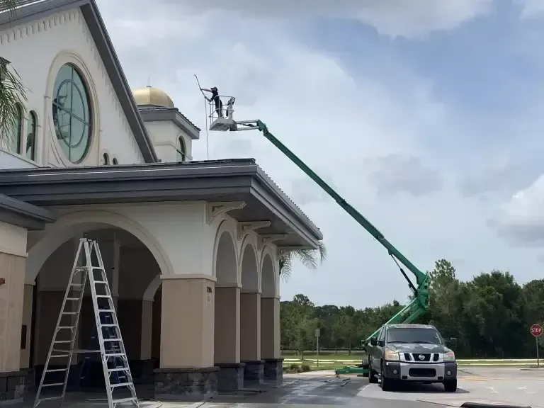 Worker on a green boom lift painting a building’s roofline beside a ladder and parked truck.