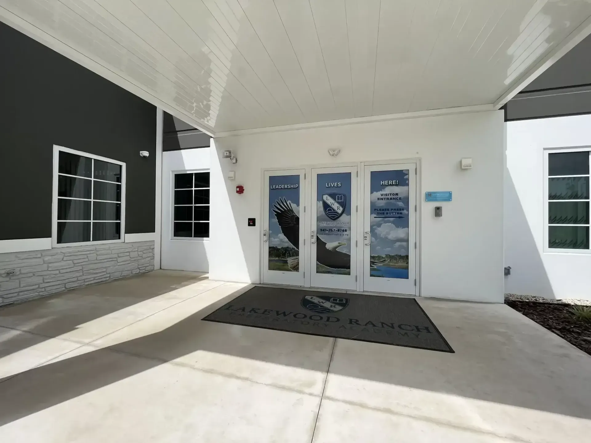 Covered building entrance with glass doors, white walls, and a black doormat under an overhang.