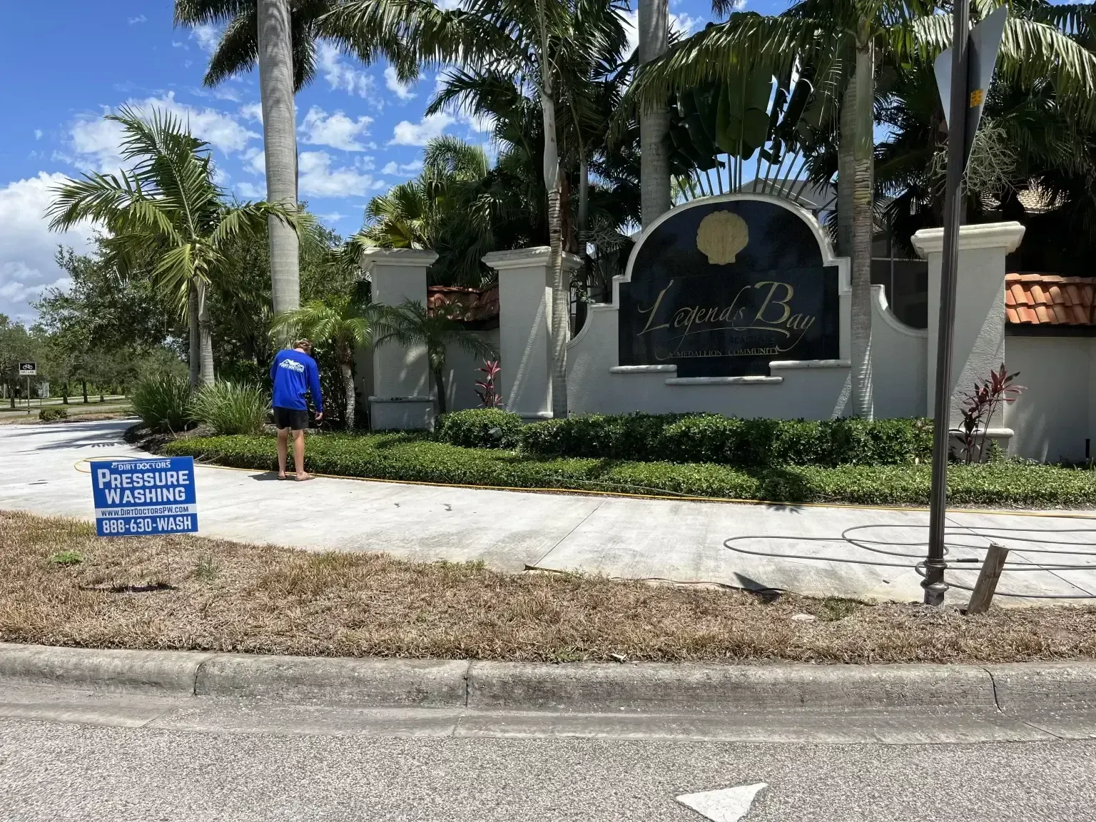 Gated community entrance with palm trees, stone sign, and a person in a blue shirt walking nearby