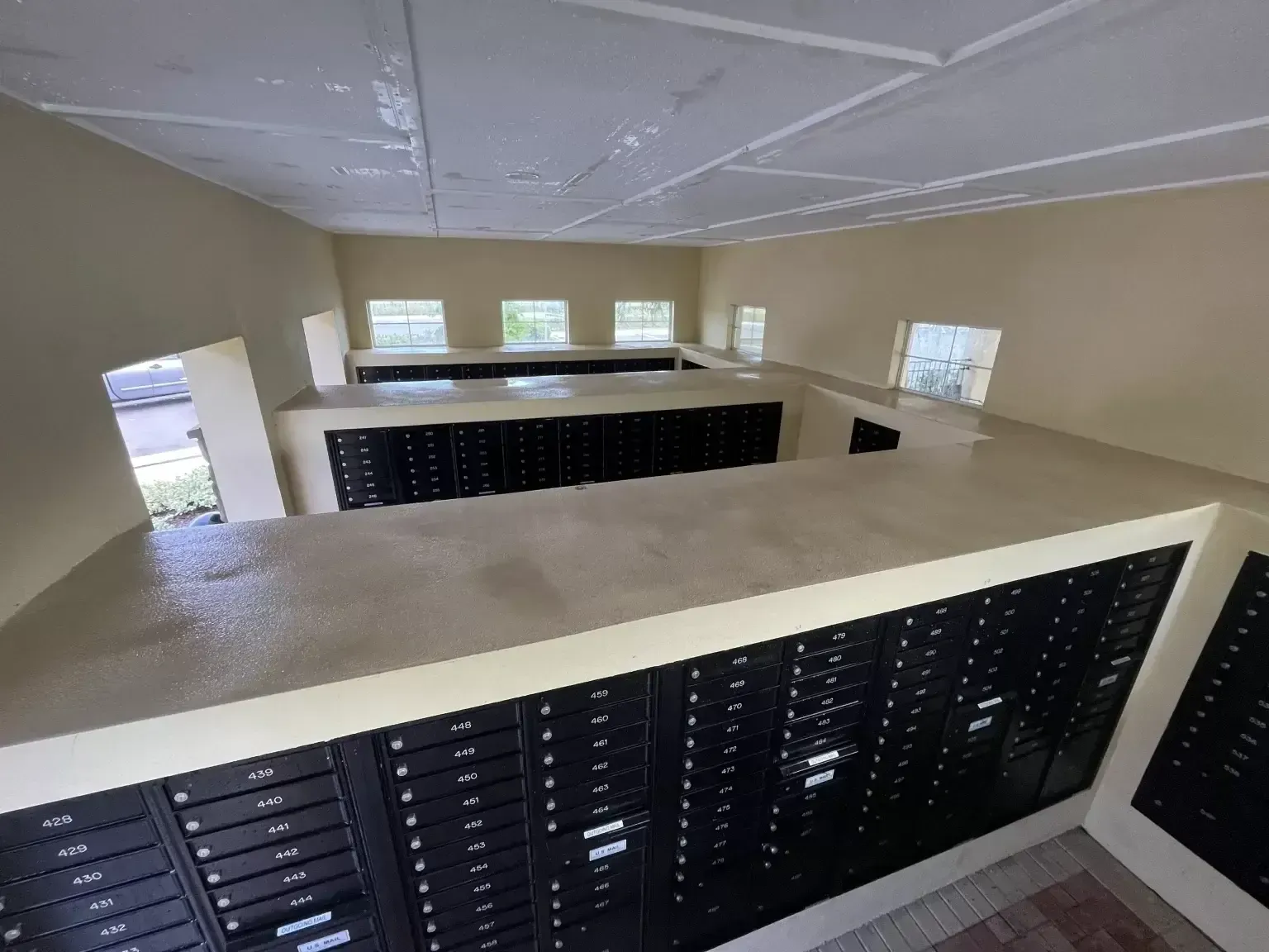 Interior of a mailroom with black mailboxes and beige walls, viewed from an upper balcony.
