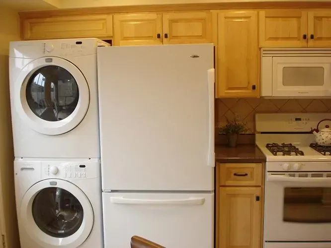 Stacked washer and dryer next to a white refrigerator in a kitchen with light wood cabinets and appliances.