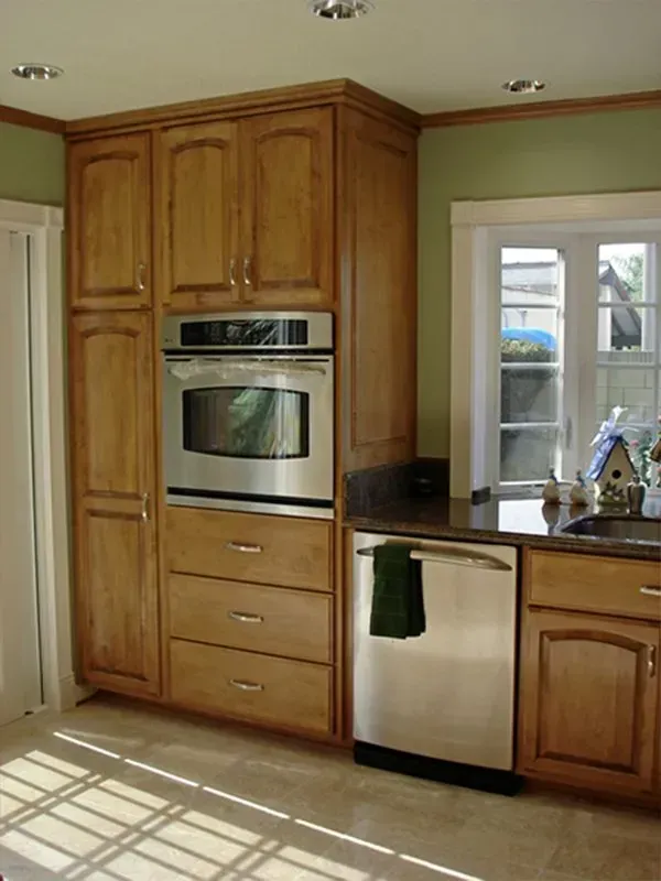 Kitchen corner with light brown cabinets, stainless steel oven, and dishwasher. Green wall with small window.