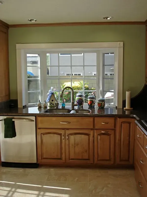 Kitchen with sink in front of a window, light wood cabinets, green walls, and stainless steel dishwasher.
