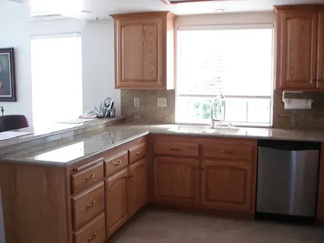 Kitchen with light wood cabinets, granite countertops, and stainless steel dishwasher. Sunlight streams in from windows.