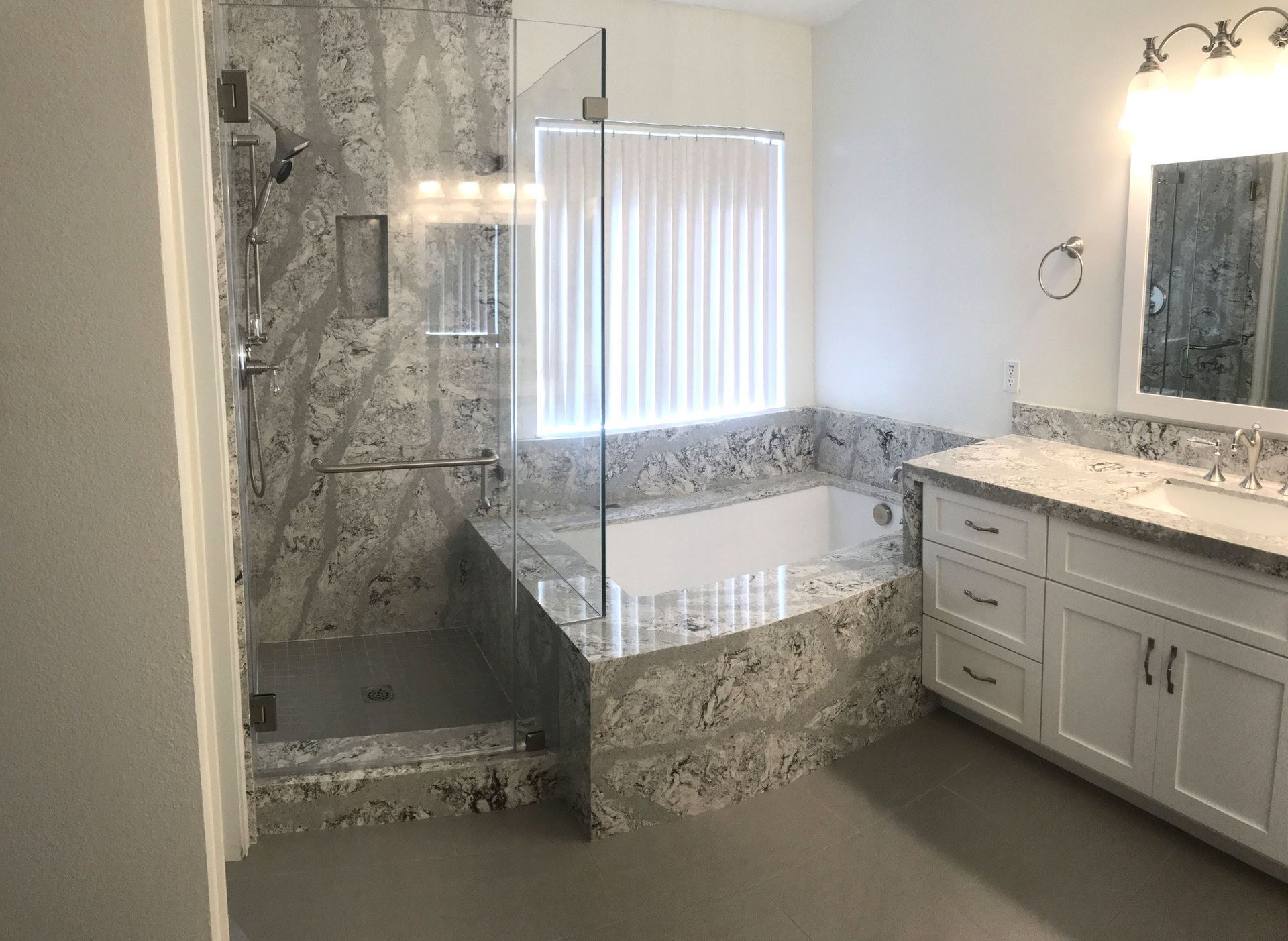 A spacious bathroom featuring a gray-patterned shower and countertop, white cabinetry, and a large window.