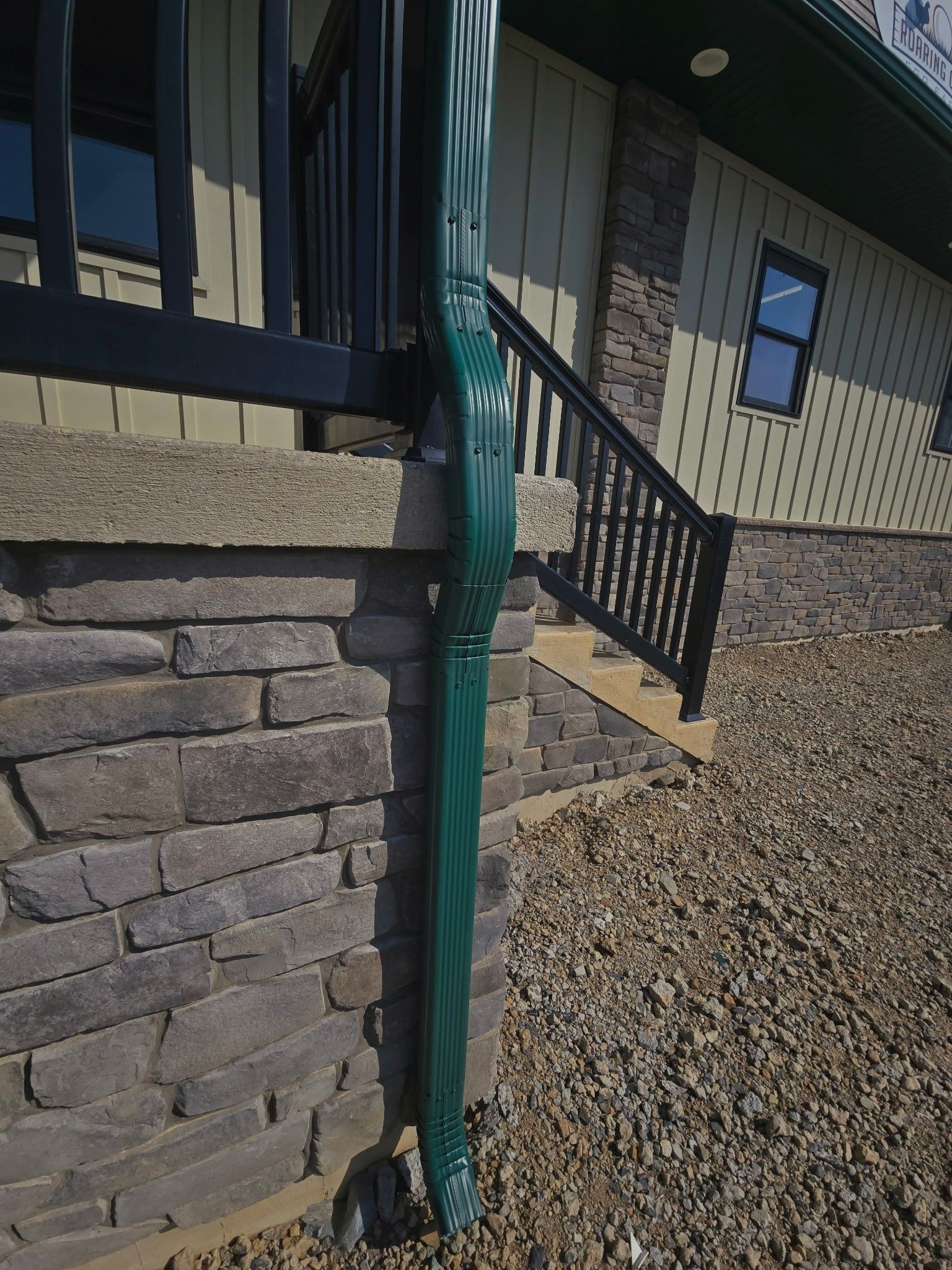 Green downspout on stone-and-siding house exterior beside gravel and stairs
