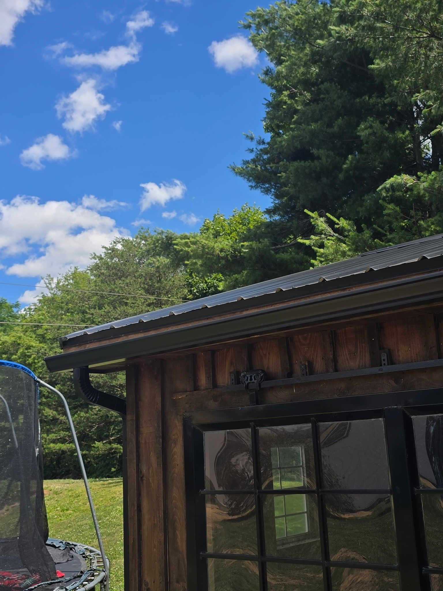 Wooden shed with black roof and window beside a trampoline, under a blue sky with trees