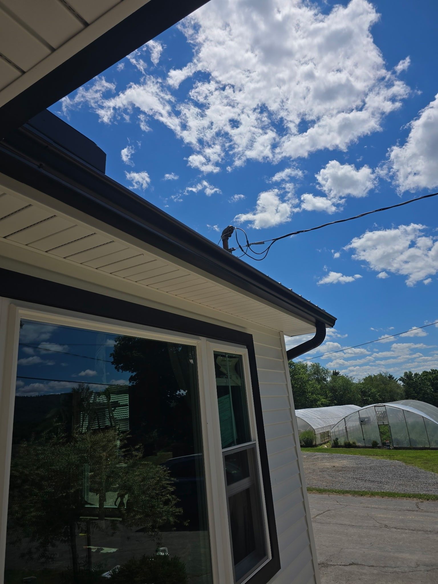 House eave and window beneath a bright blue sky with clouds, power lines overhead, and white greenhouses nearby