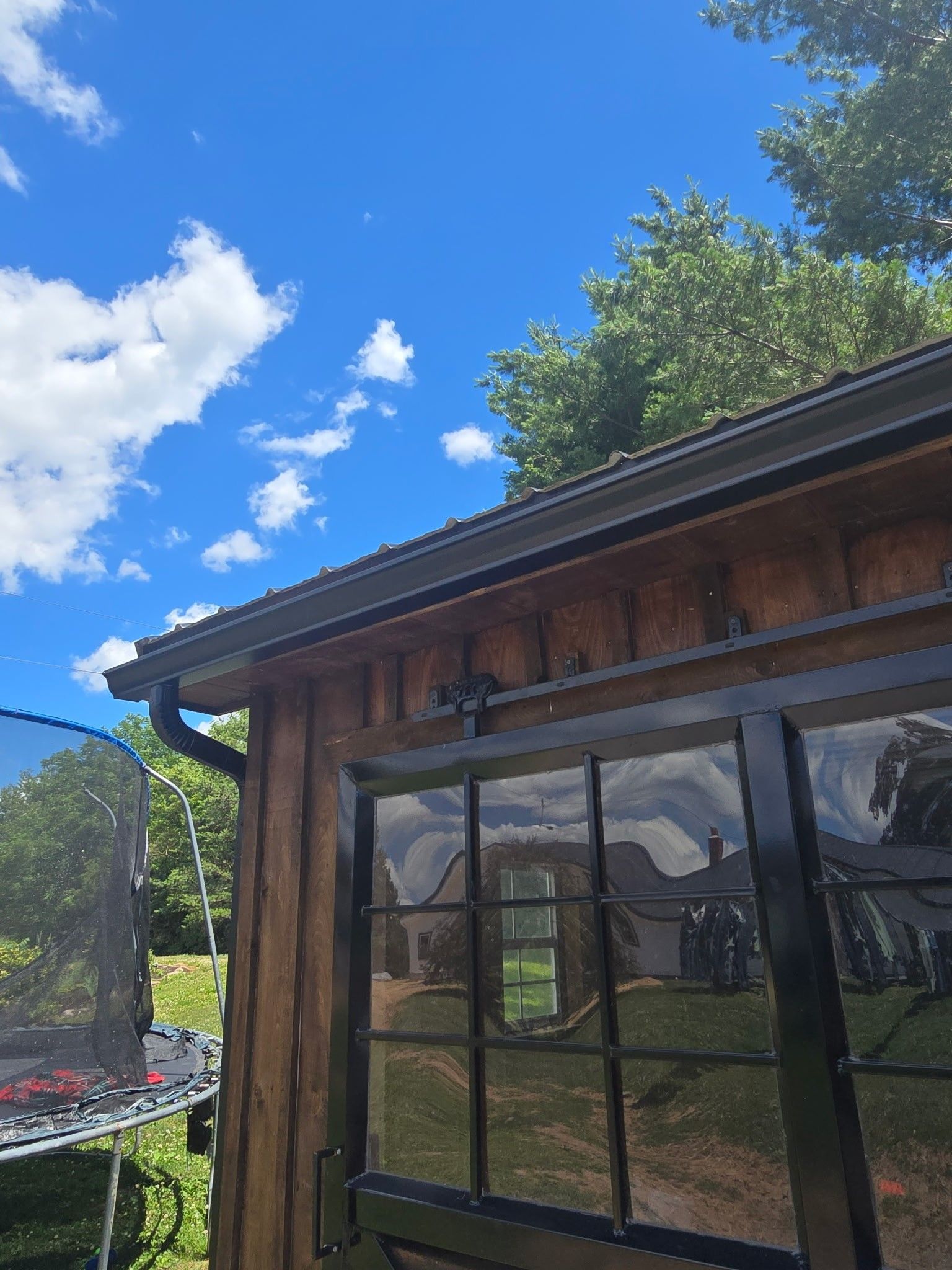 Wooden house exterior with large windows under a bright blue sky, with a trampoline beside it