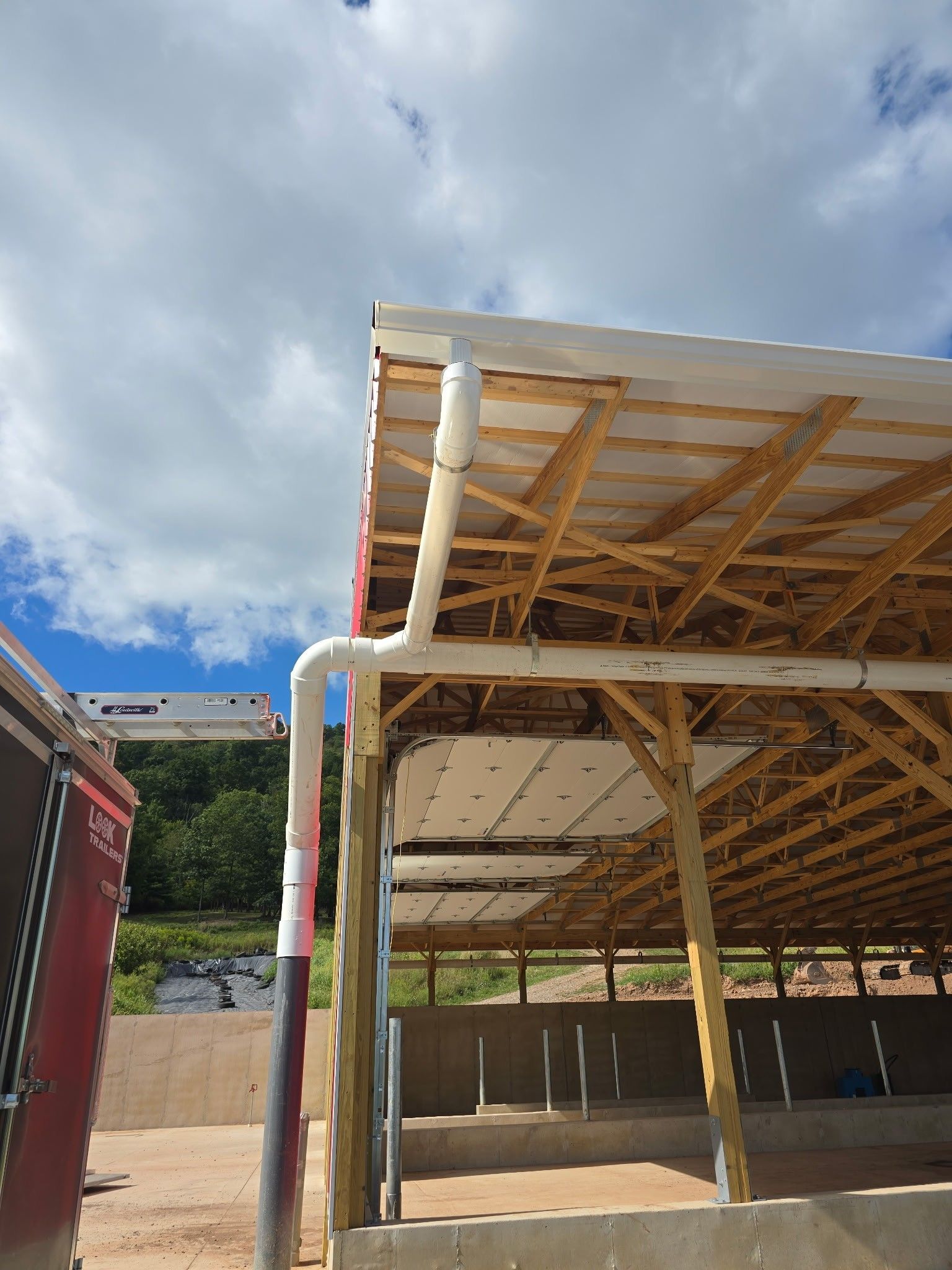 Open-sided industrial shed with wooden roof trusses under a cloudy sky.