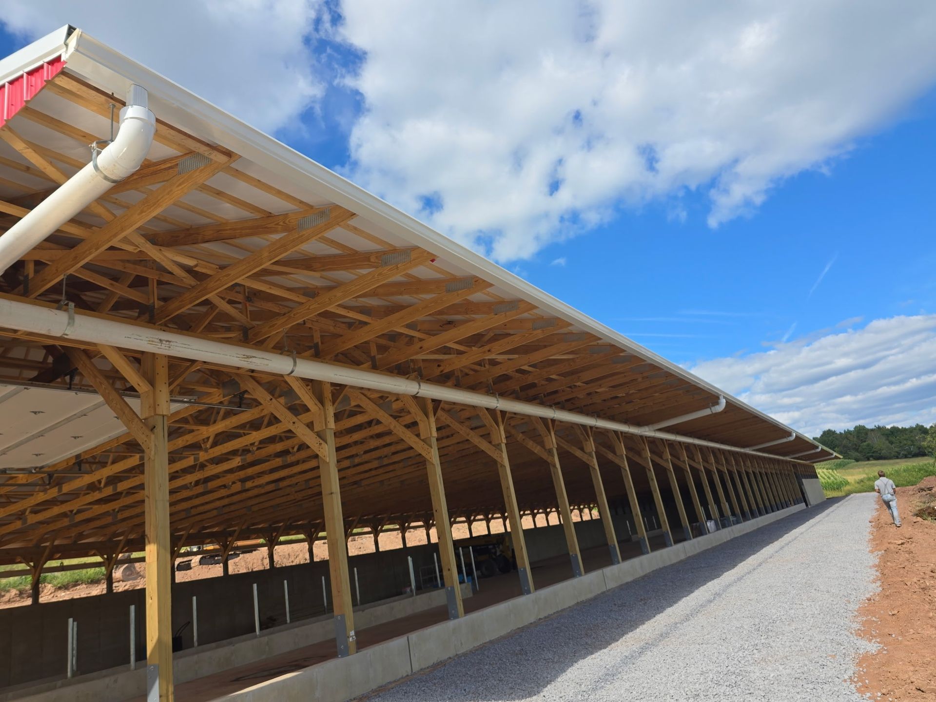 Open-sided wooden farm shelter with a metal roof along a gravel path under a blue sky