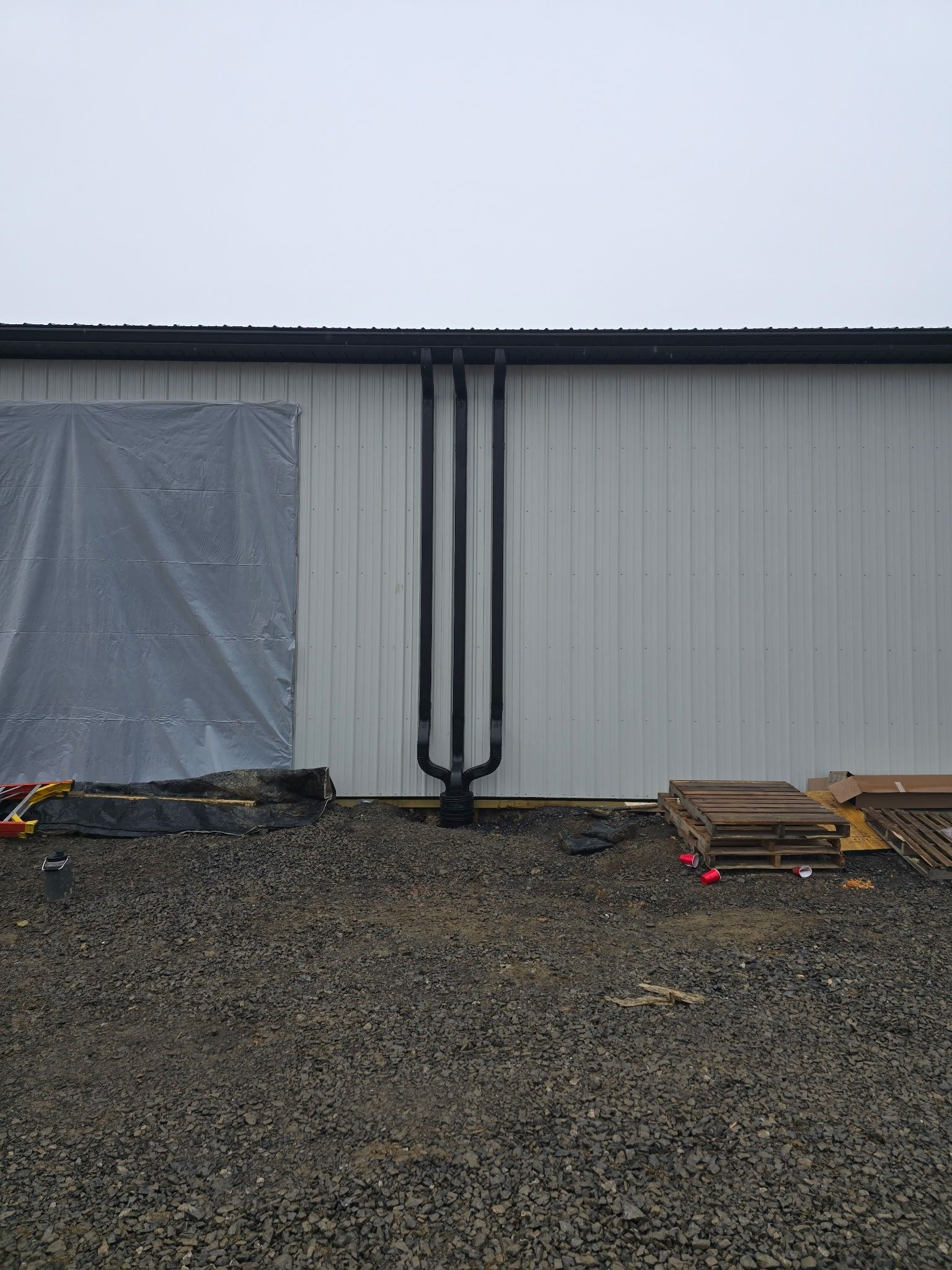 Industrial building wall with a covered doorway, black downspout, gravel lot, and stacked pallets on the right