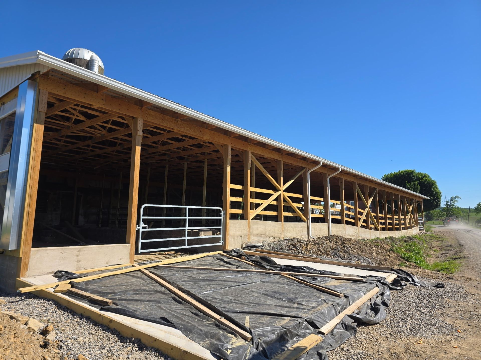 Open-sided barn with a corrugated roof, wooden framing, and a gravel lot under a clear blue sky