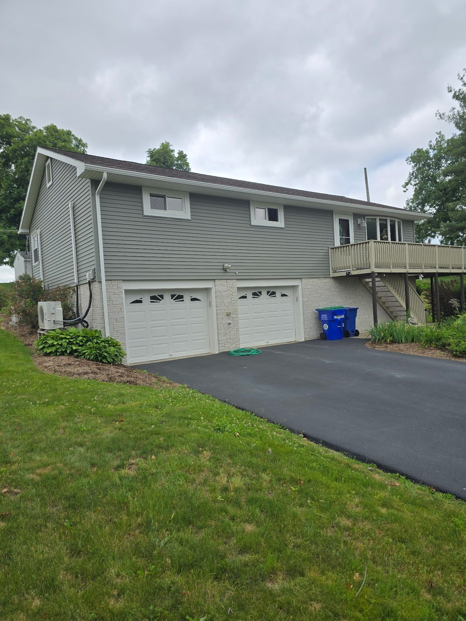 Gray two-story house with double garage, sloped driveway, and blue recycling bins under cloudy sky