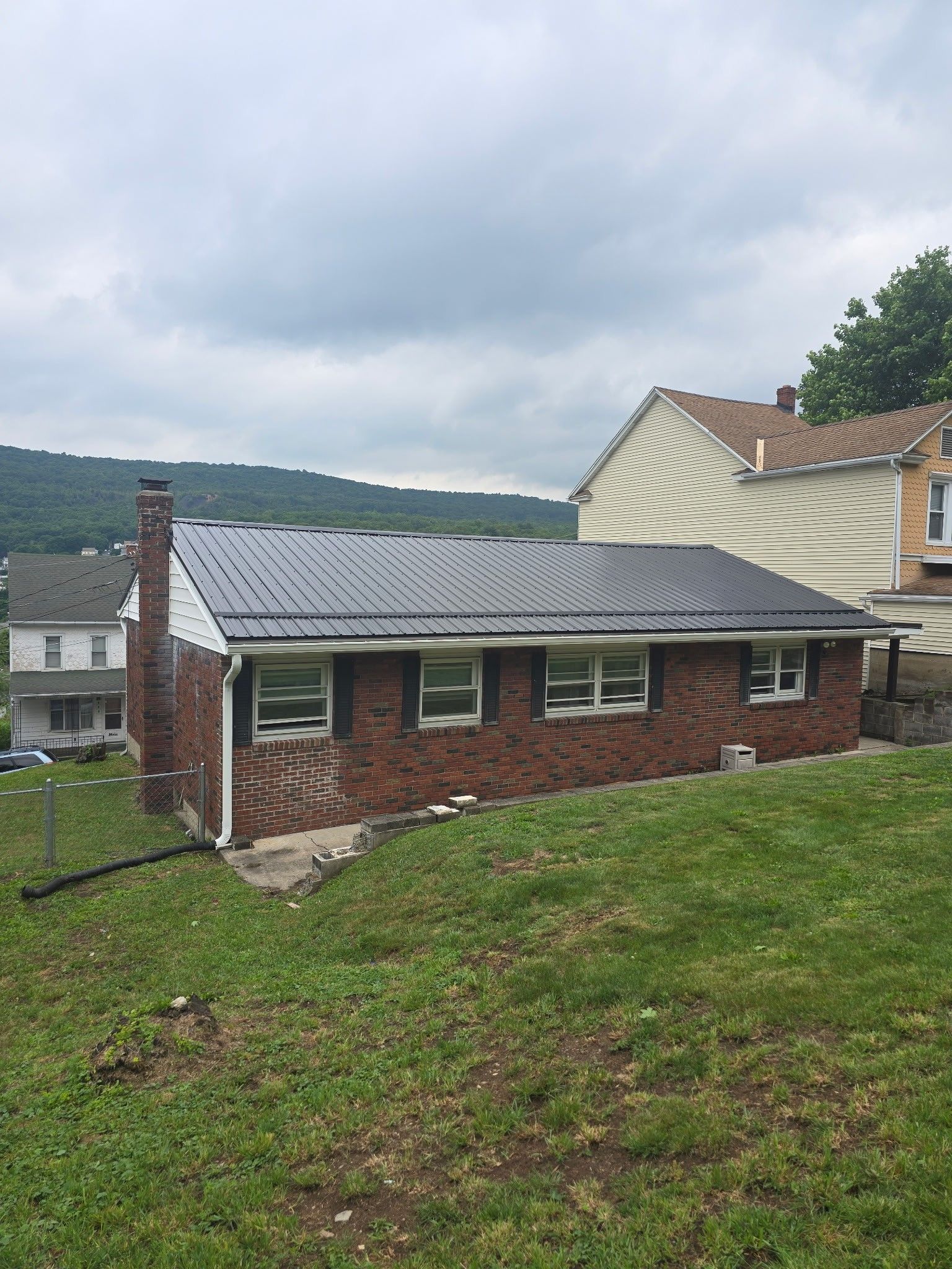 Brick house with a gray metal roof on a grassy hillside under cloudy skies