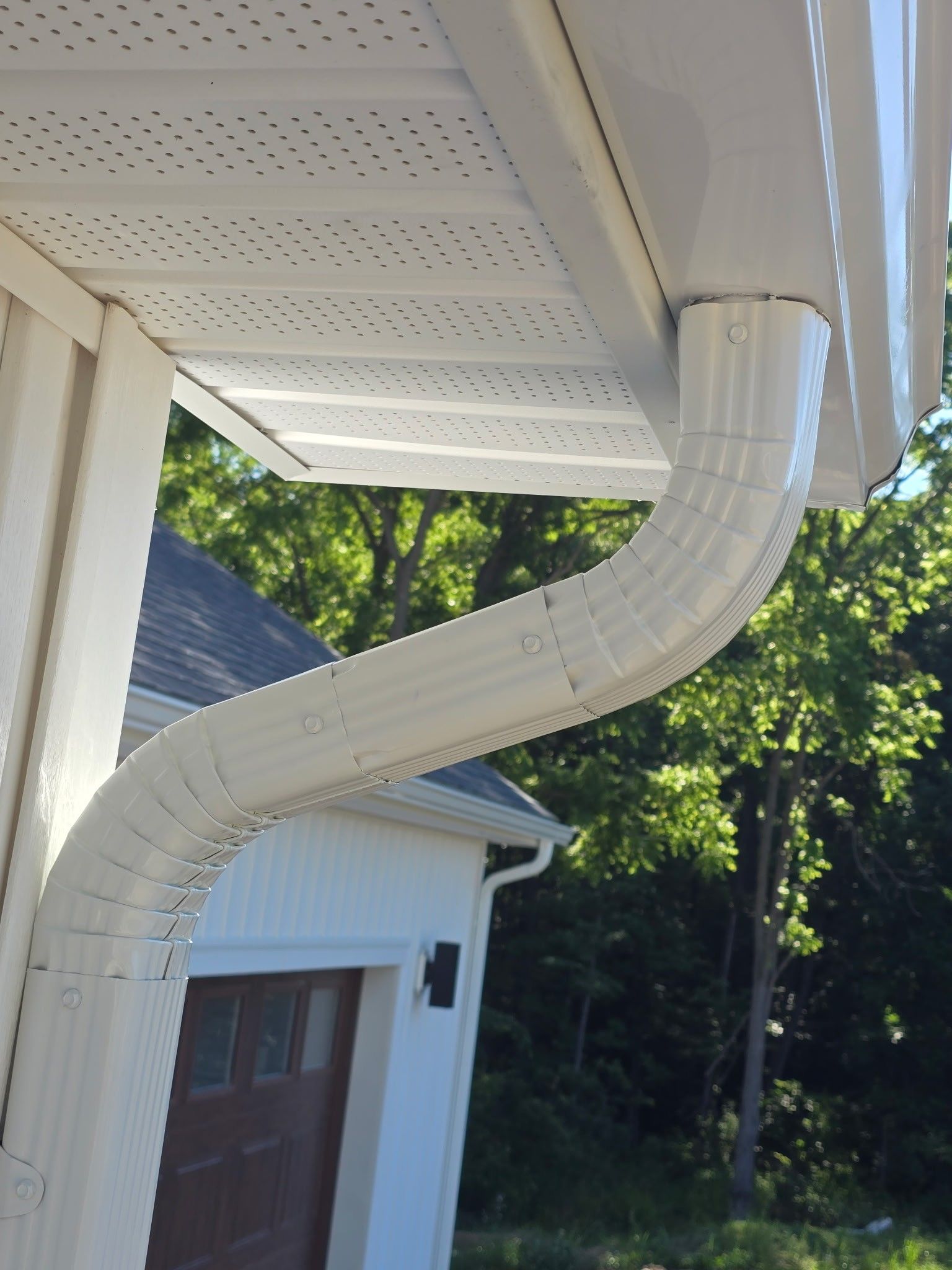 White gutter downspout curving from roof eaves beside a house, with trees in the background