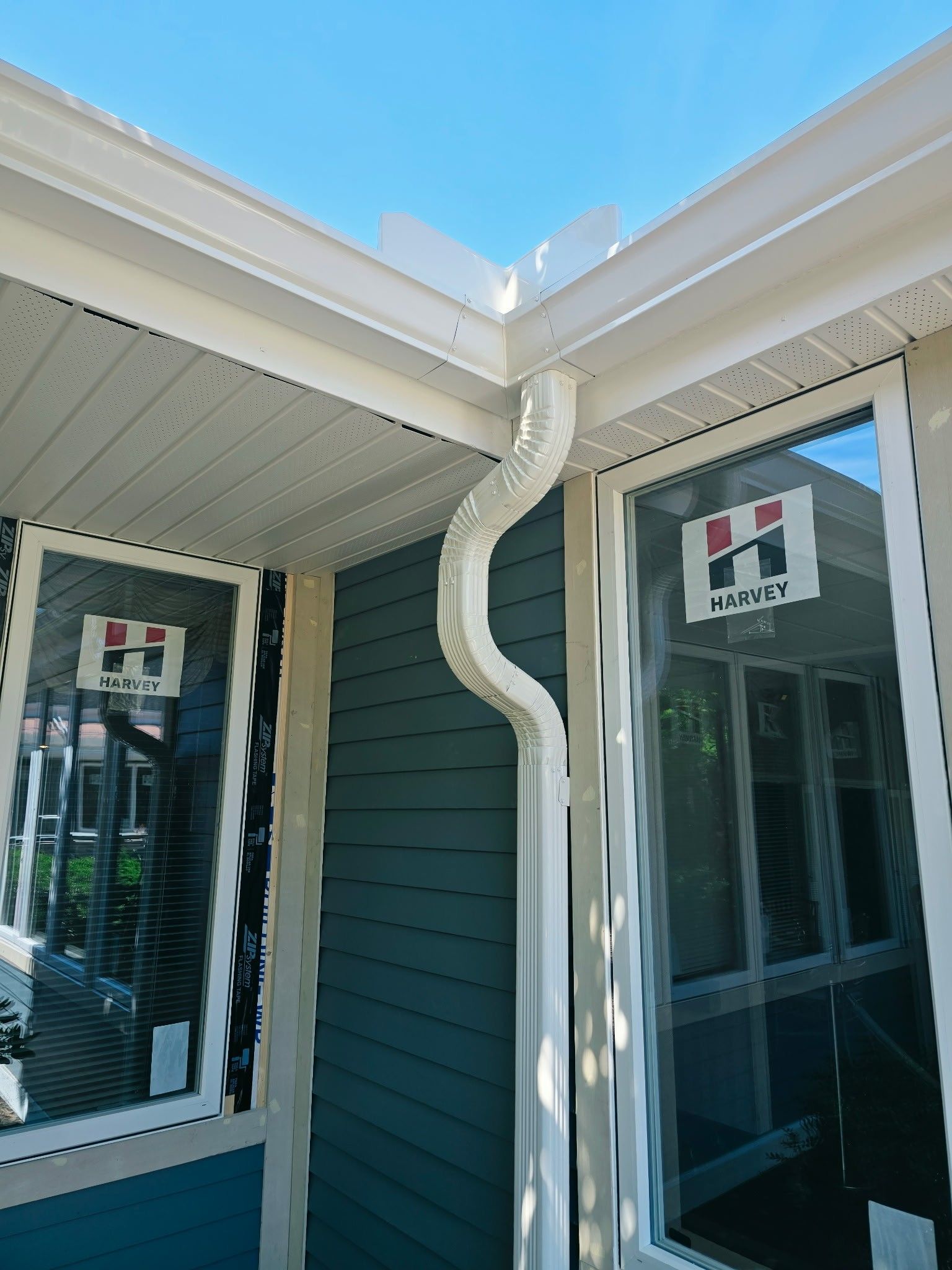 Blue house siding and white eaves with a gutter downspout between two windows under a bright sky