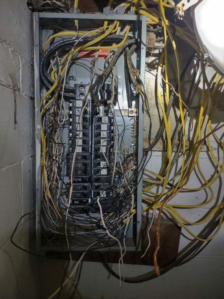 Open electrical panel with tangled yellow and black wires in a basement setting.