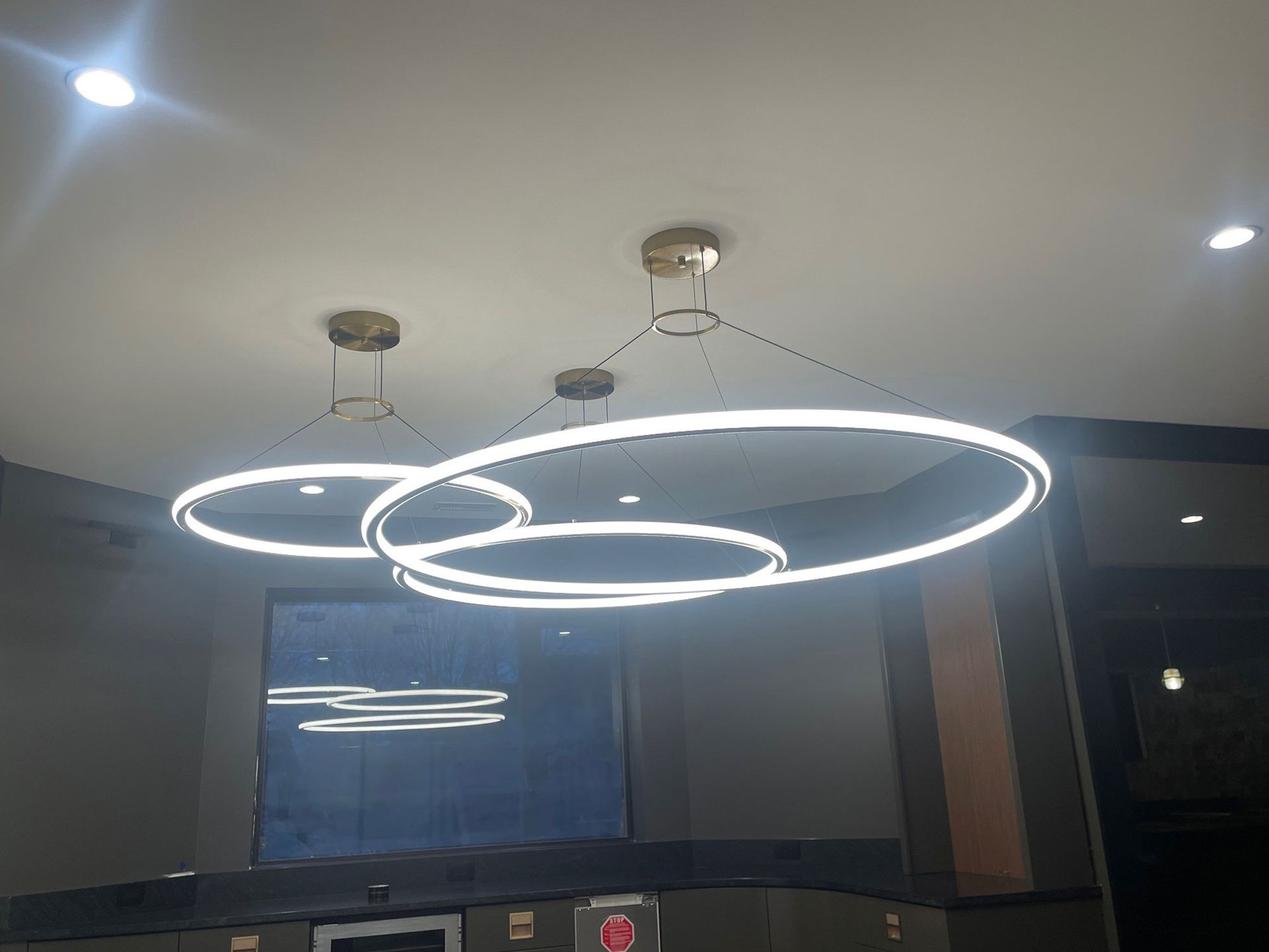 Three modern, glowing ring lights hanging from a ceiling, reflecting in a window.