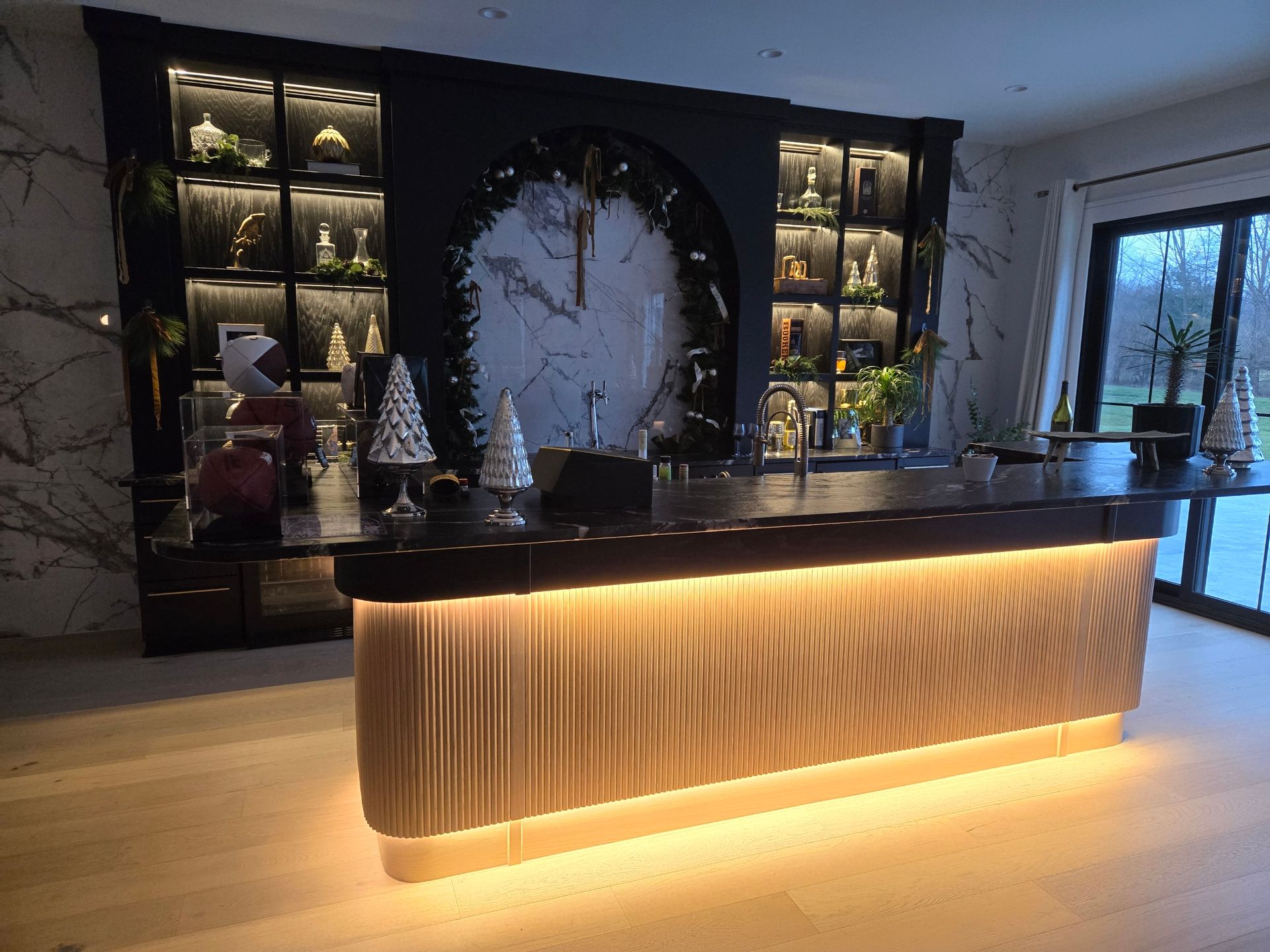 A bar with a backlit textured facade, a large clock, and lit shelves displaying items, with a window in the background.