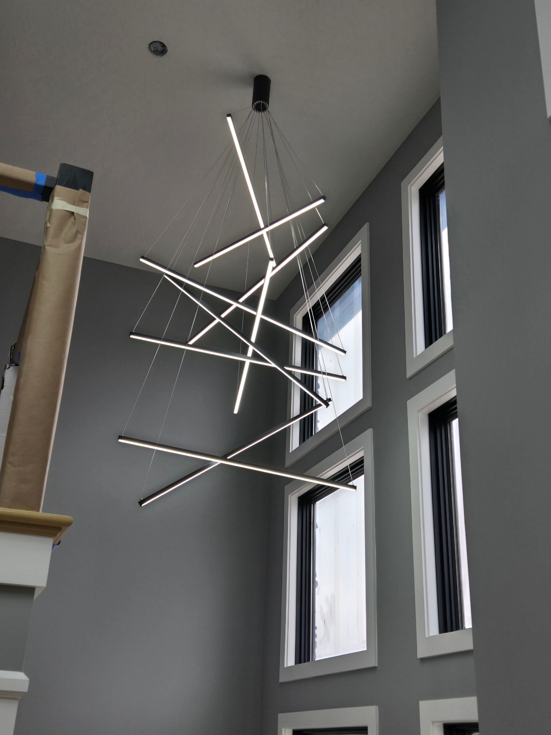 Modern chandelier with intersecting light bars hangs in a gray-walled room with tall windows.