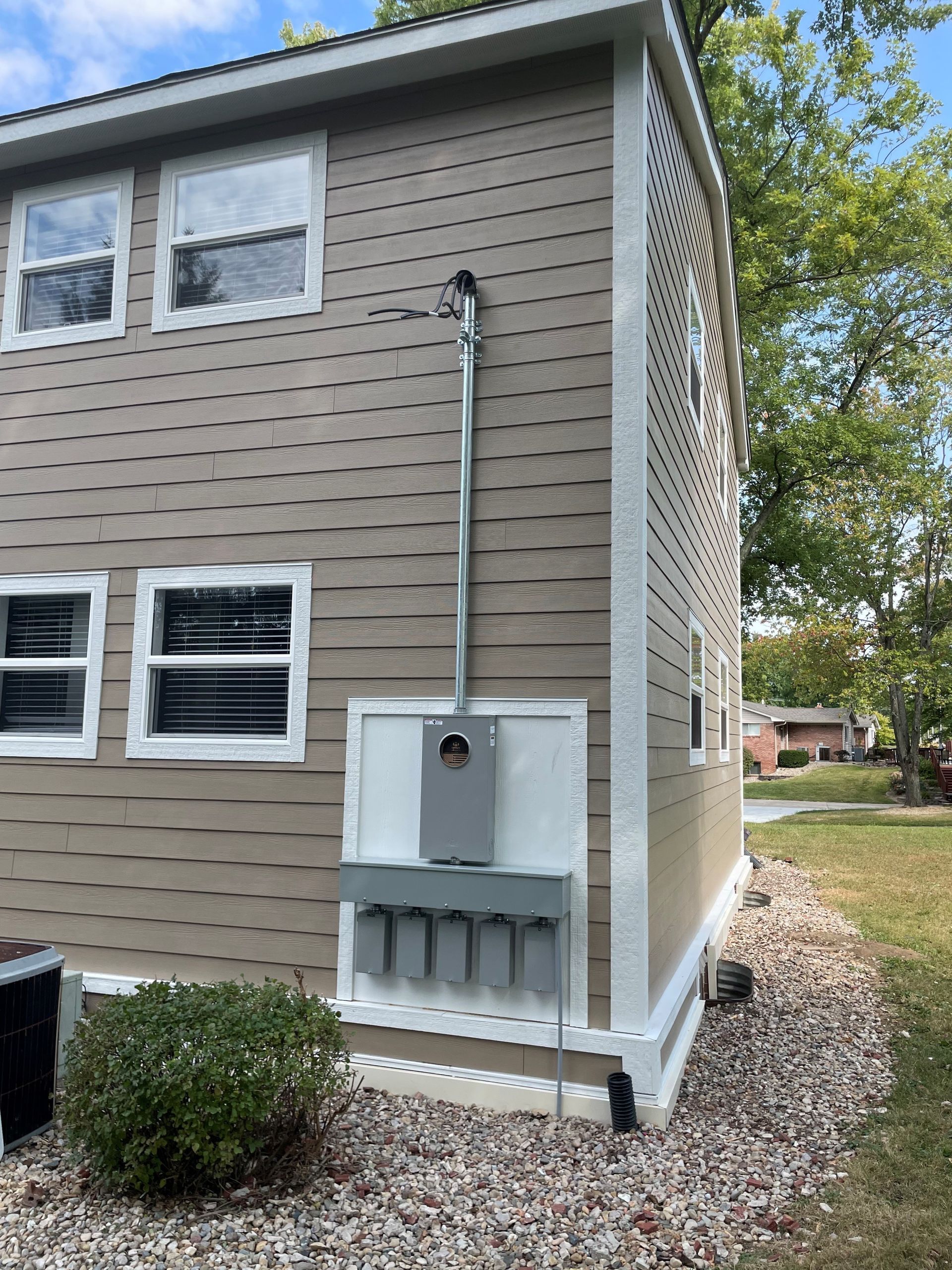 Exterior wall of a house with electrical panel and conduit, mounted on beige siding, gravel ground.