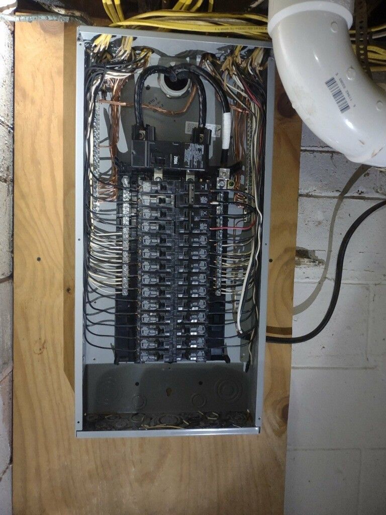 Electrical panel with numerous circuit breakers, mounted on a wood board in a basement setting.