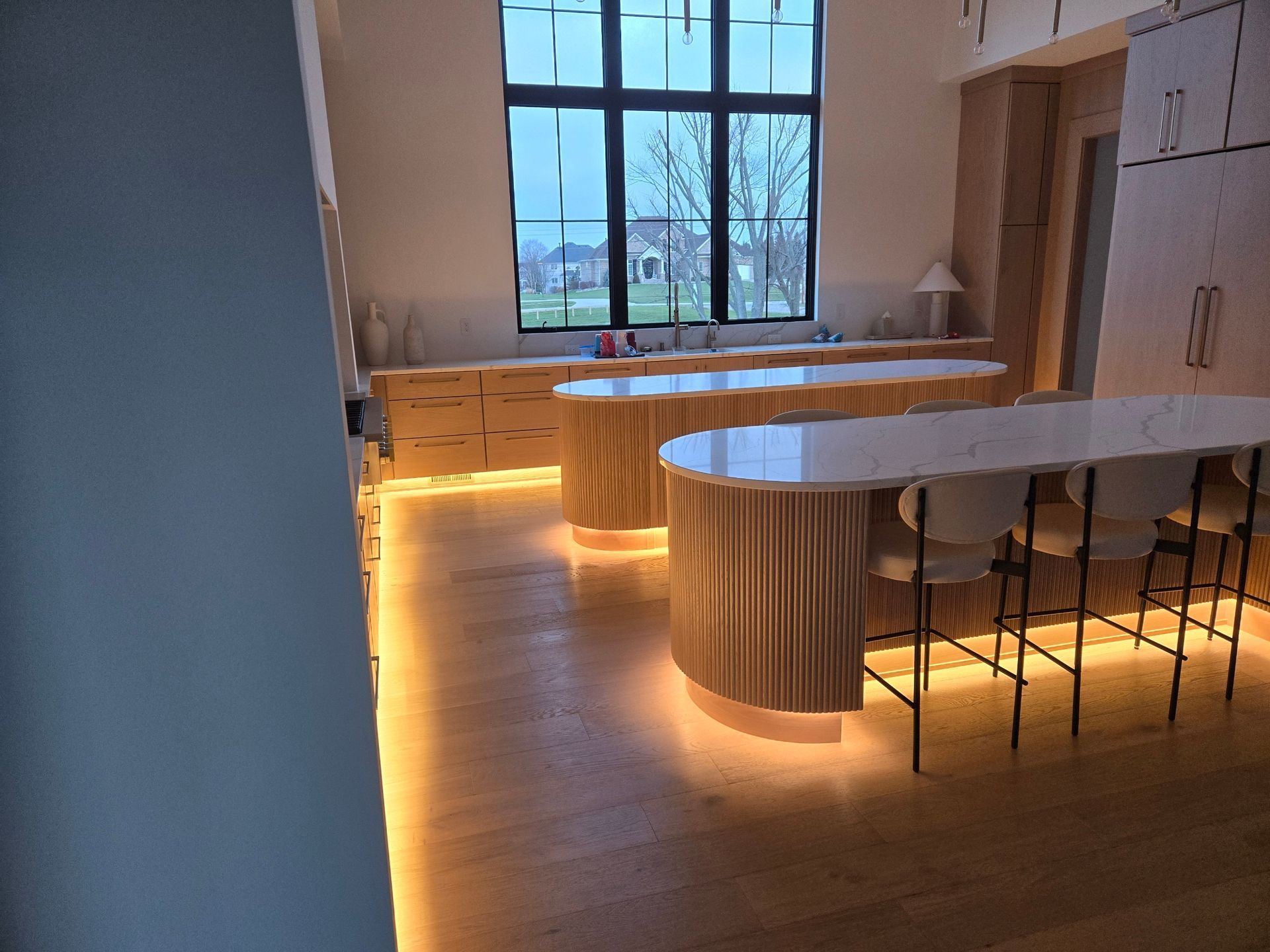 Kitchen with wood flooring, two light-up curved islands, white countertops, large window, and bar stools.