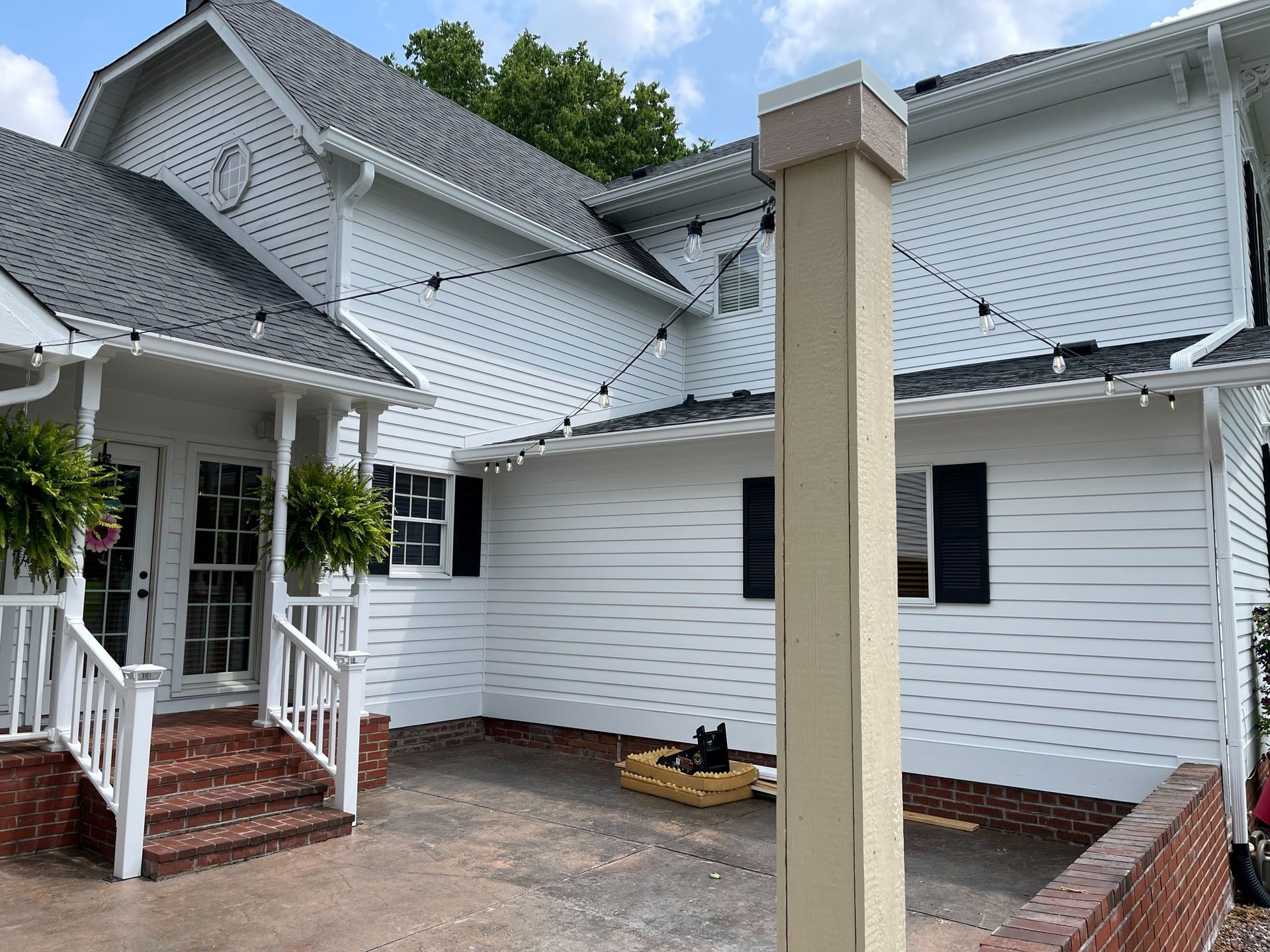 White house exterior with black shutters, gray roof, and brick patio; string lights.