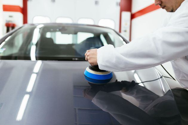 Person polishing a black car hood in an auto shop with a handheld buffer.
