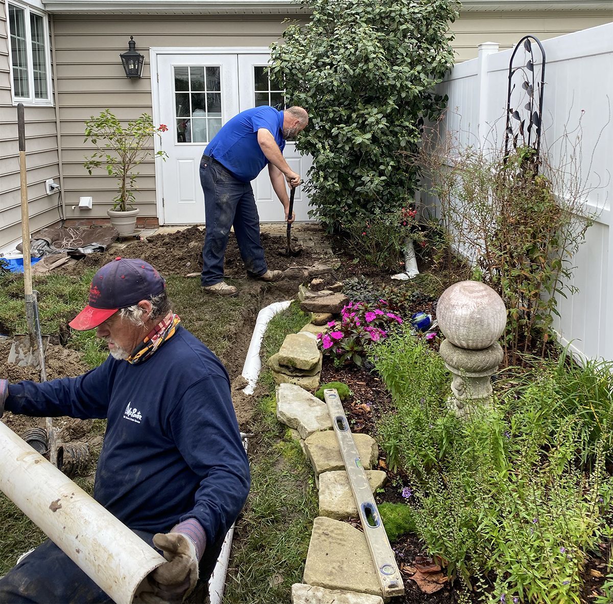 Two people gardening beside a house, digging and carrying soil among flower beds and shrubs.