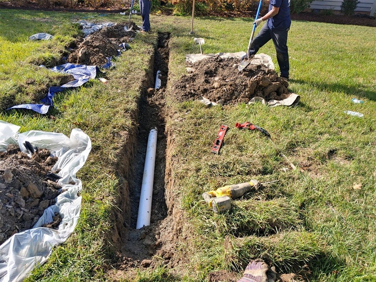 Workers digging a trench beside a white pipe in a grassy yard, with sod piles and plastic sheeting.