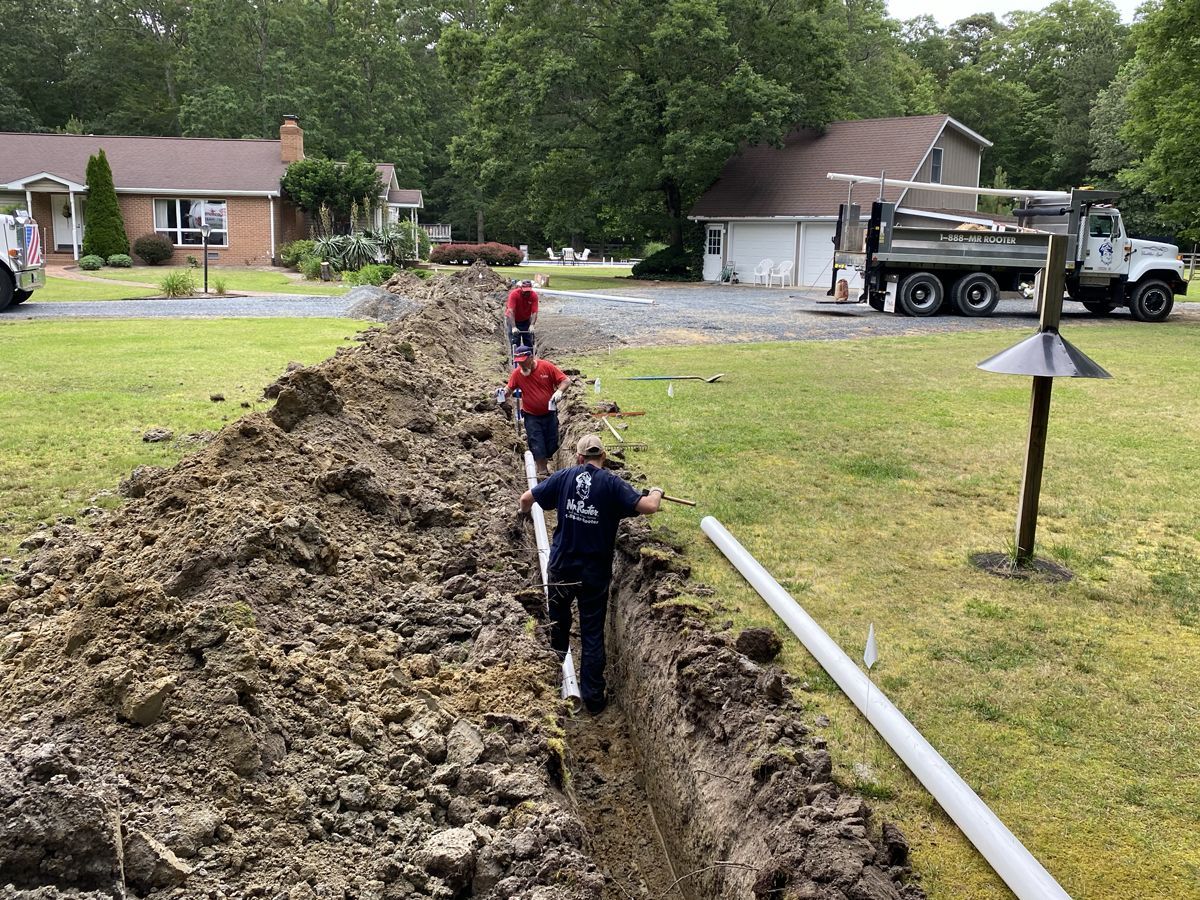 Workers in a deep trench laying pipe beside a muddy dirt pile in a suburban yard.