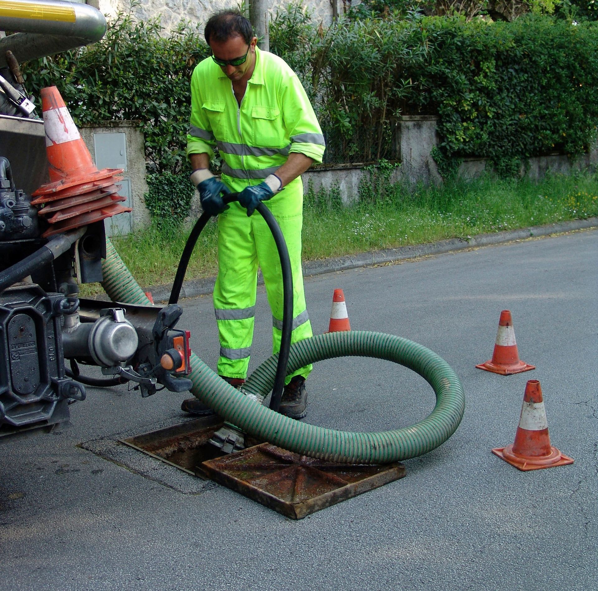 Worker in neon green safety gear handling a large hose beside a street repair truck and traffic cones