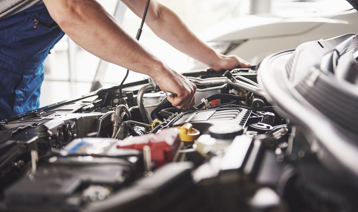 Mechanic working on a car engine under the hood. Blue work overalls, inside a garage.