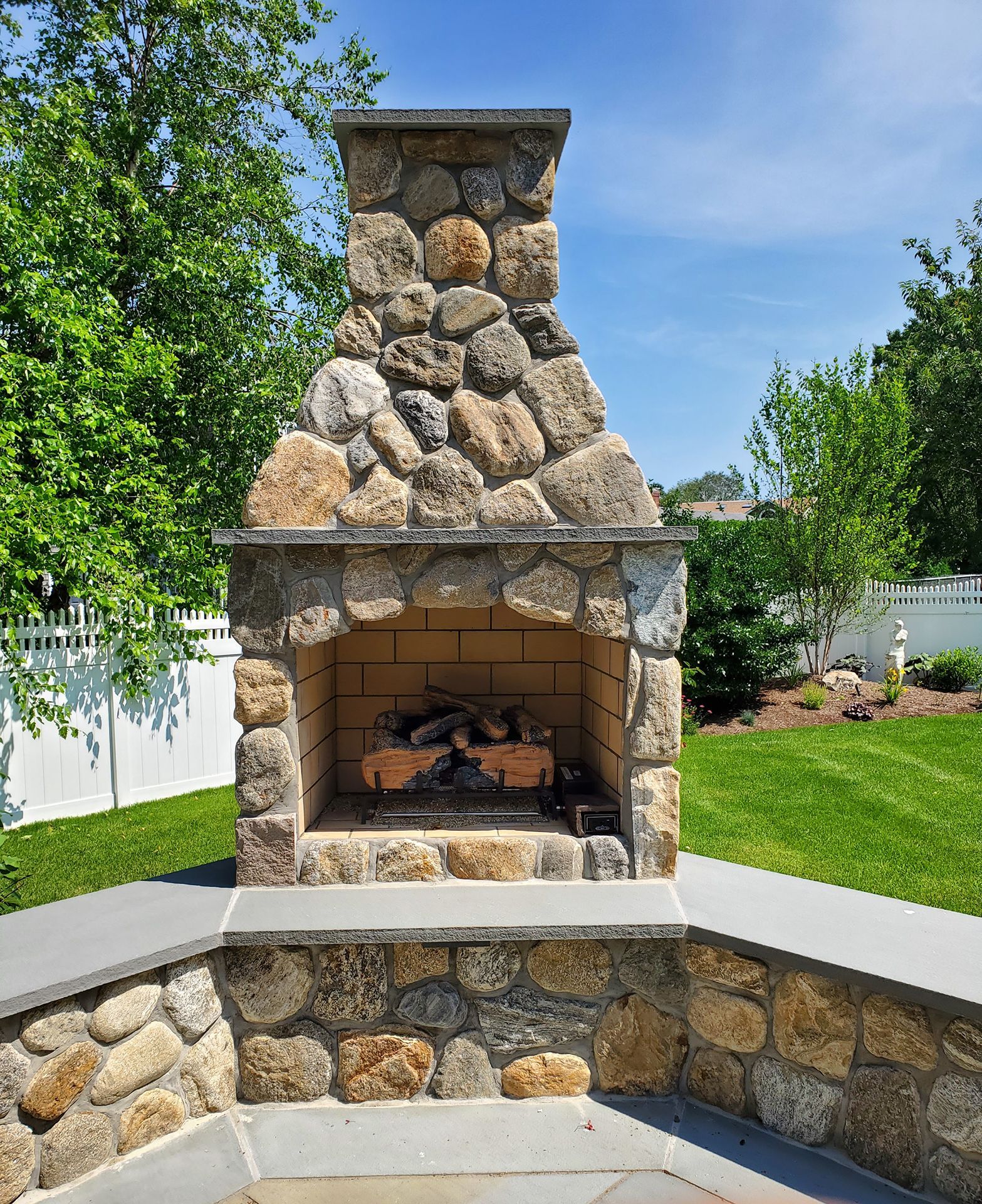 A stone fireplace in a backyard with a white fence in the background