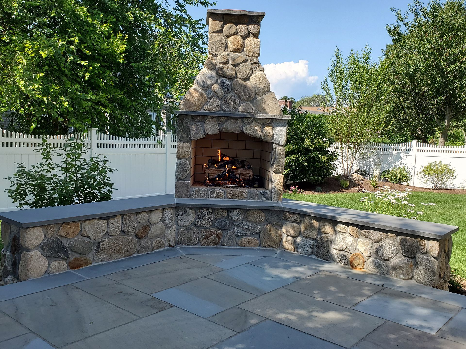 A stone fireplace in a backyard with a white fence in the background