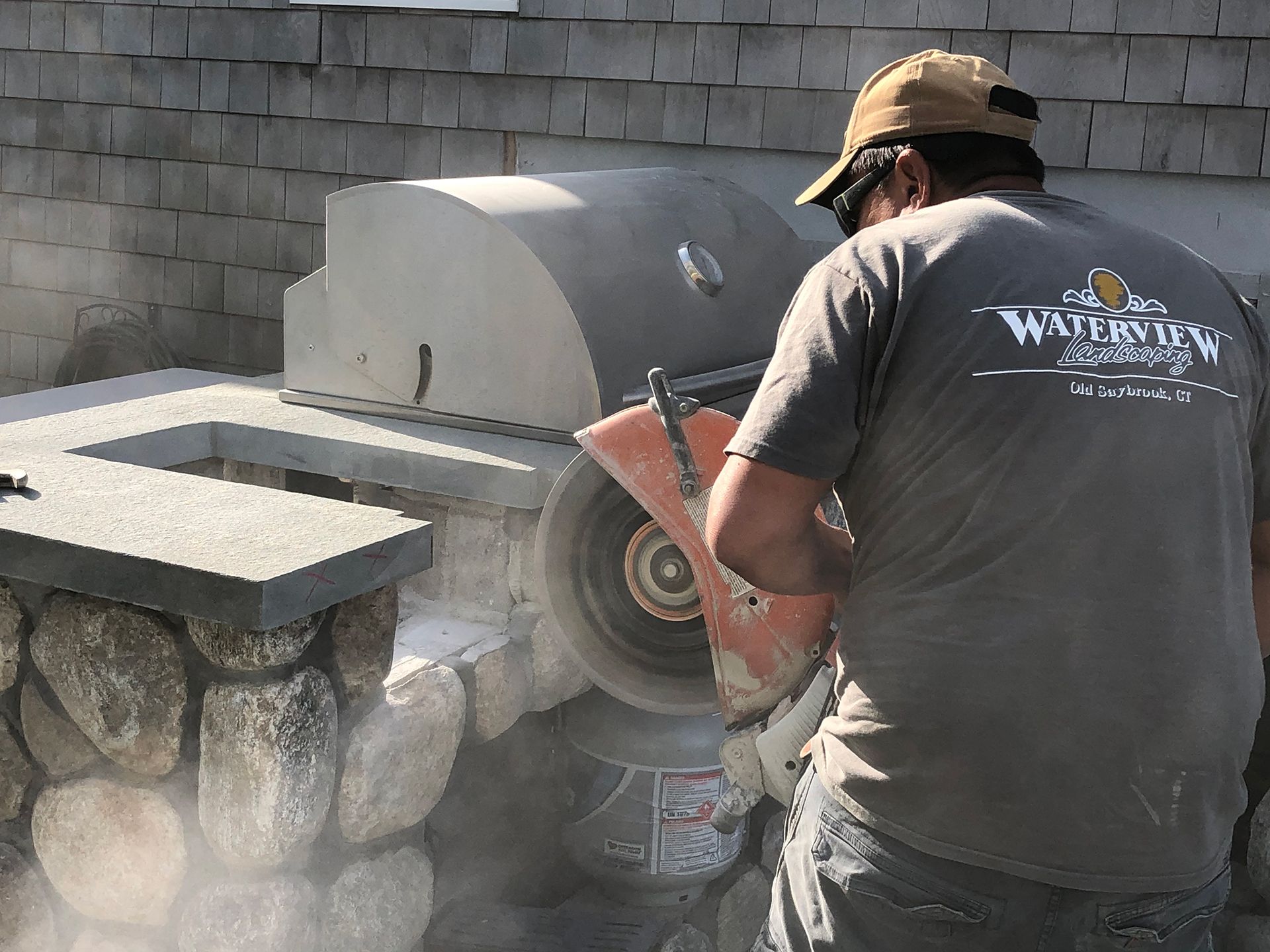 A man wearing a waterbury shirt is cutting a stone wall with a circular saw.