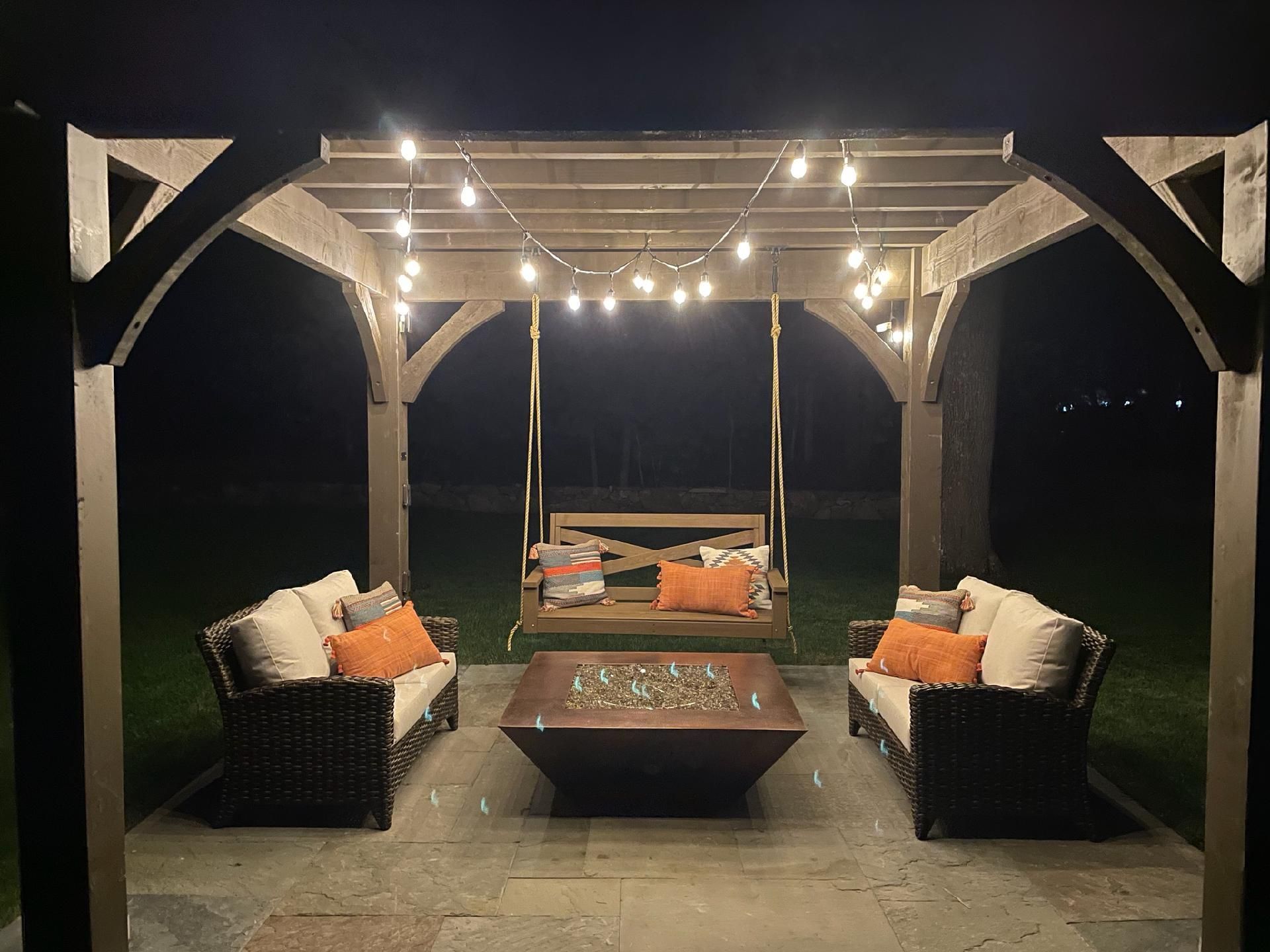A pergola with a swing, couch, coffee table, and fire pit at night.
