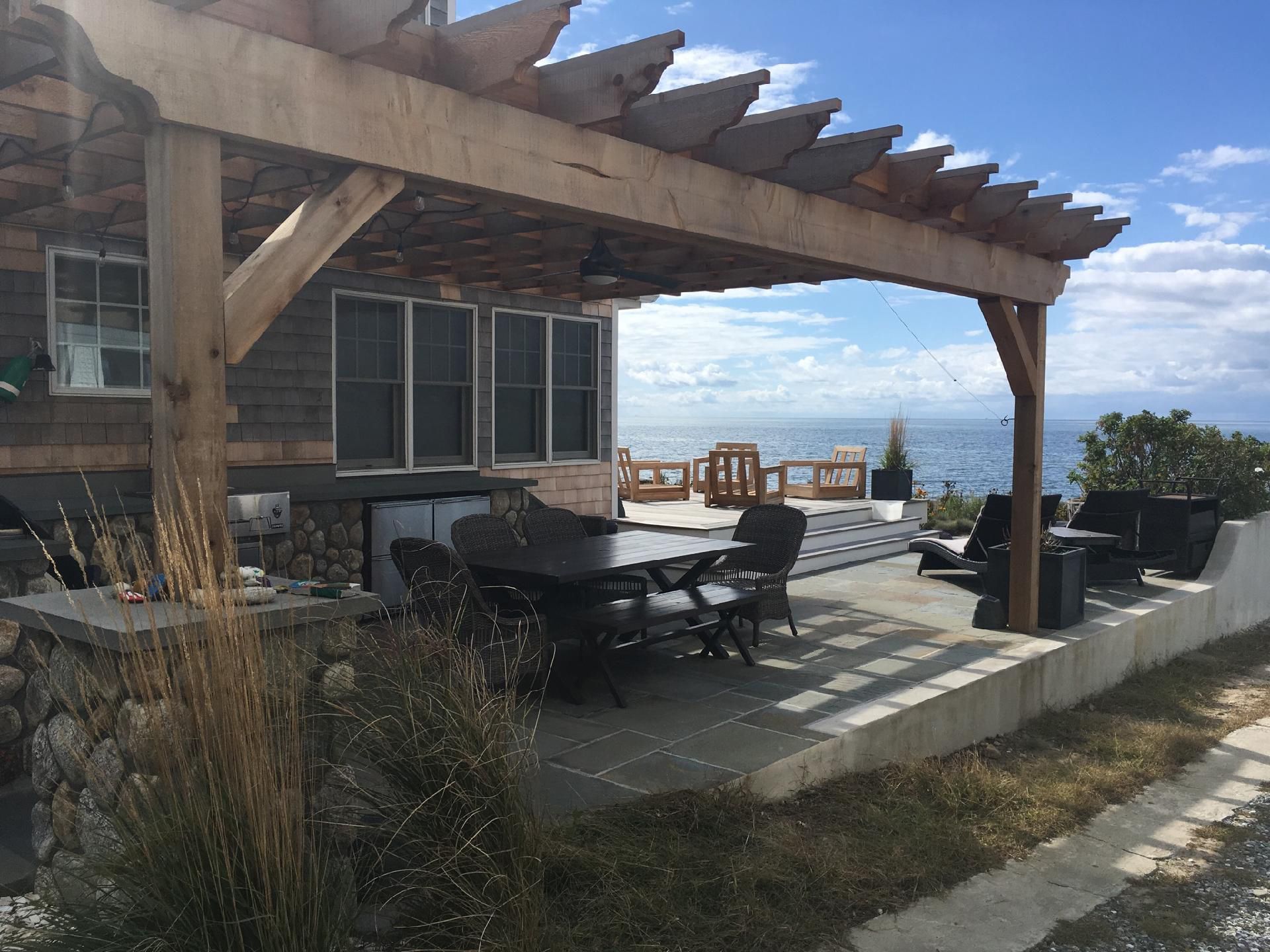 A patio with a table and chairs under a pergola overlooking the ocean