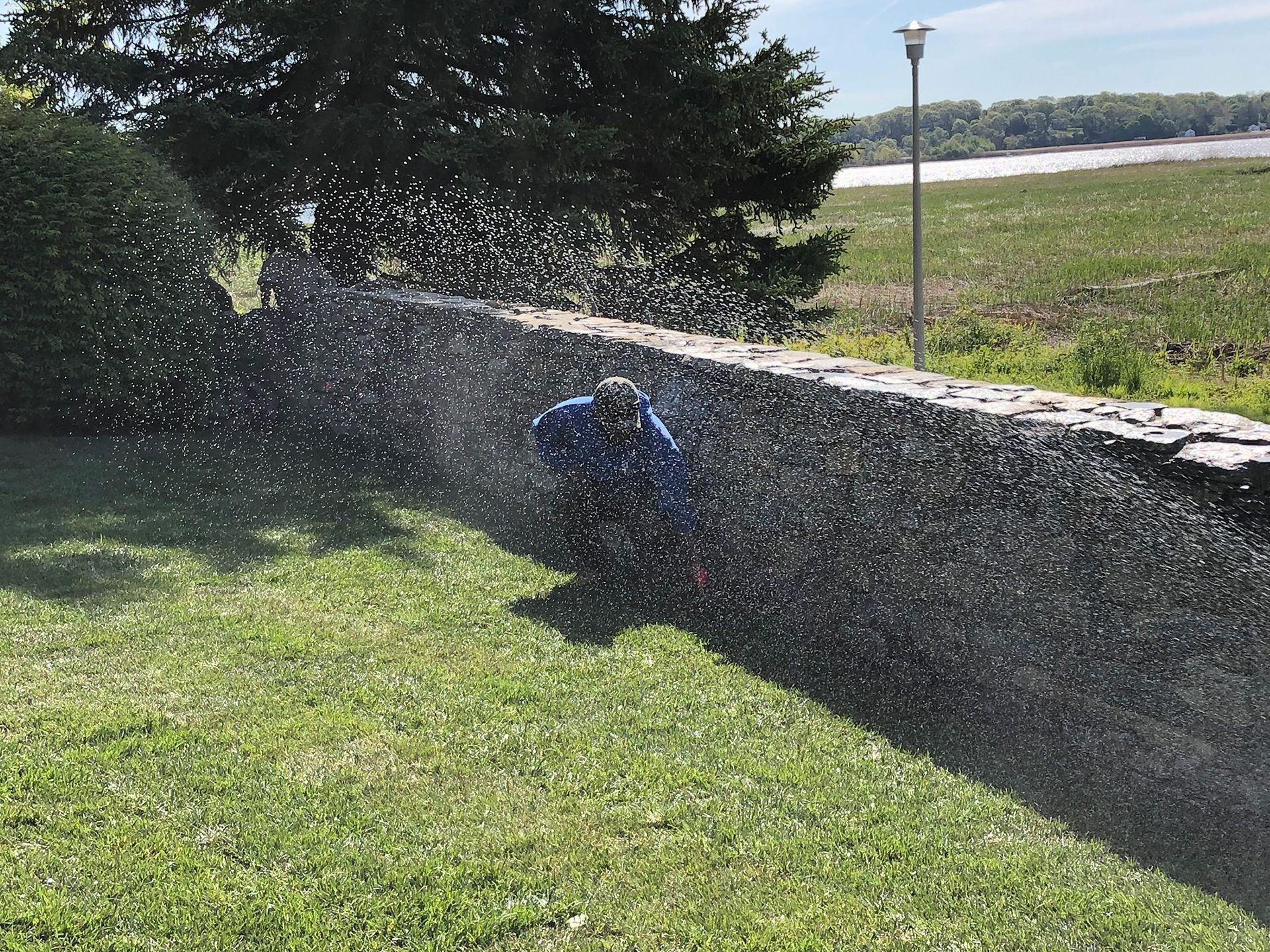 A person standing next to a stone wall spraying water on the grass