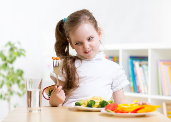 A little girl is sitting at a table eating vegetables with a fork