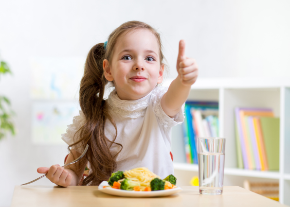 A little girl is sitting at a table with a plate of food and giving a thumbs up