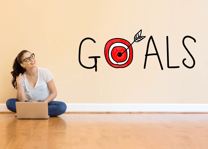 A woman is sitting on the floor with a laptop in front of a wall with the word goals written on it