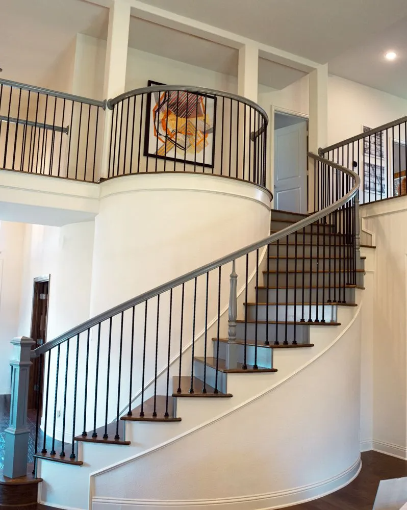 Curved staircase with metal railings, ascending to a second-floor balcony. White walls, artwork, bright interior.